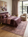 5' x 8' Rectangular Brown Beige Distressed Modern Abstract Area Rug displayed in a bedroom beneath a bed with coordinated mauve bedding.