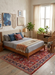 7' x 10' Rectangle Red & Navy Floral Traditional Washable Area Rug placed in a cozy bedroom with a wooden bed frame, neutral bedding, and natural light from a large window.