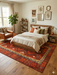 7' x 9' rectangular red geometric tribal southwestern area rug with bold diamond and zigzag patterns in deep red, rust, beige, and charcoal tones, bordered by repeating tribal motifs; shown styled in a cozy bedroom and home office with warm wood furniture and neutral decor.