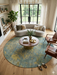 8' Round Blue & Gold Distressed Floral Traditional Washable Area Rug styled in a living room with a curved sofa, coffee table, and natural light from large windows.